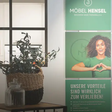 Pflanze im Korb vor zwei grünen Möbel Hensel Postern mit Logo, Frau in grünem Shirt mit Herzgeste und Text Unsere Vorteile sind zum Verlieben.